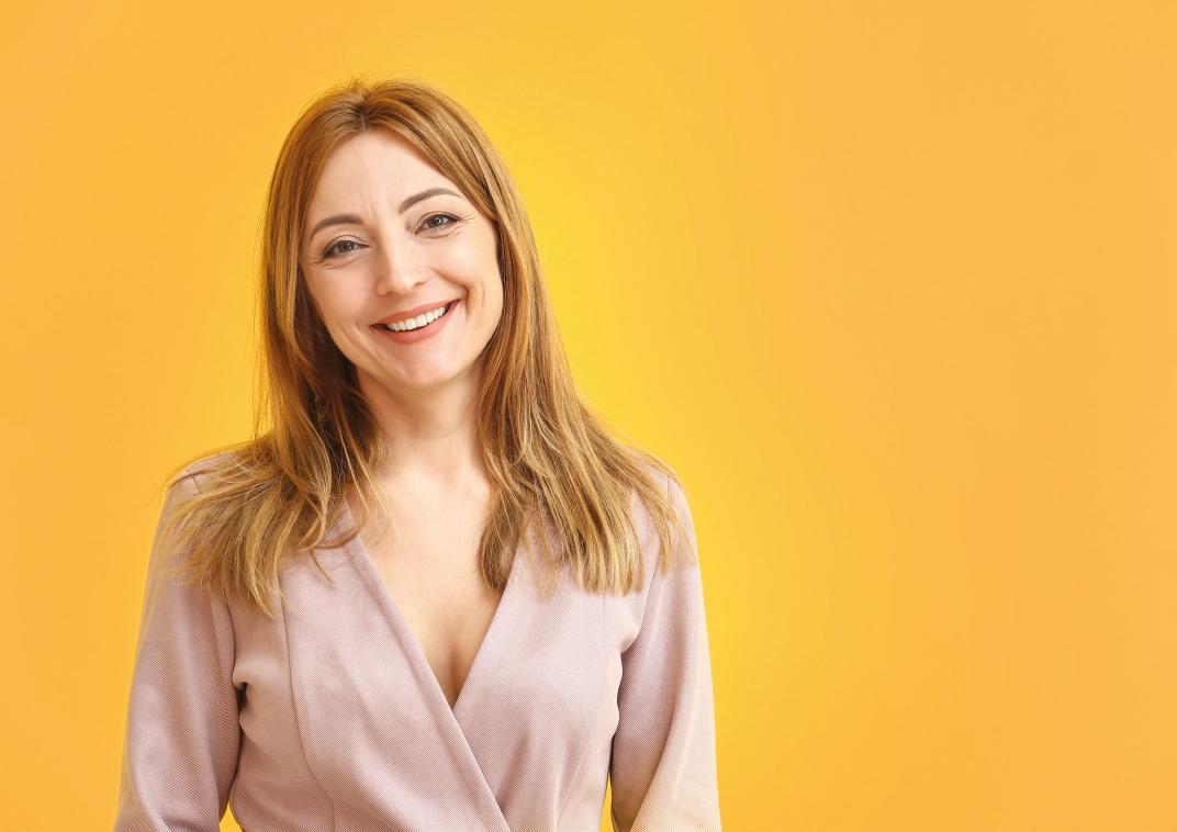 Vrouw met lang, lichtbruin haar glimlacht in een lichte blouse tegen een gele achtergrond.