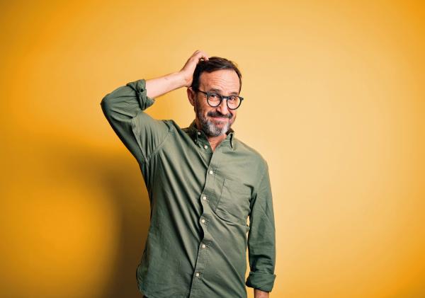 Deze afbeelding toont een man met een bril en een groene overhemd staat voor een felgele achtergrond.